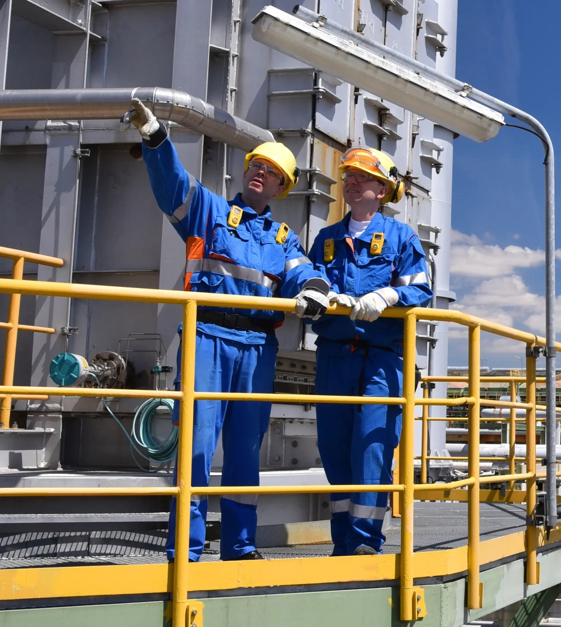 Workers in hard hats oversee industrial processes on a sunny refinery platform, ensuring safe and efficient operations